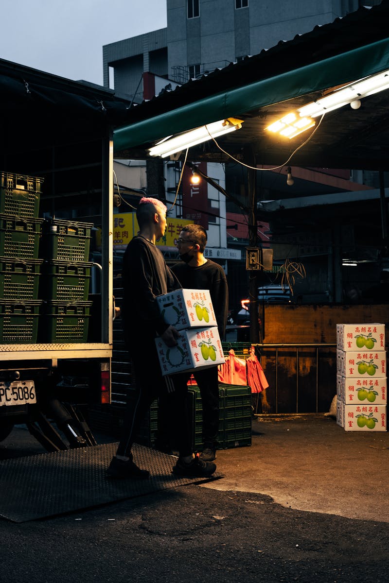 Workers unloading boxes from a delivery truck at an urban market during dusk.